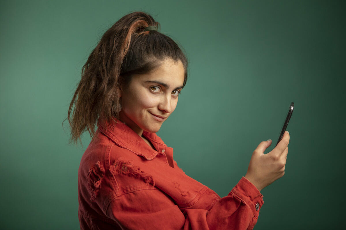 Junge Frau mit Smartphone in der Hand, die kokett in die Kamera guckt und vor einem dunkelgrünen Hintergrund steht. © Westend61 / Getty Images