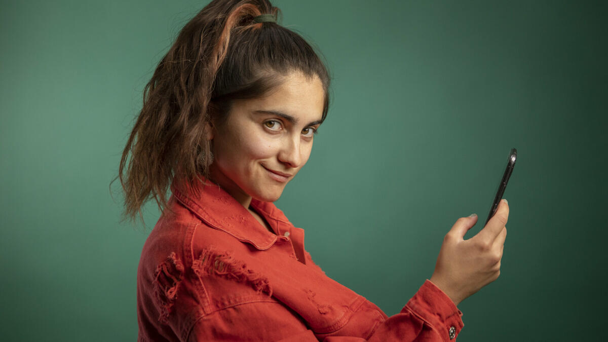 Junge Frau mit Smartphone in der Hand, die kokett in die Kamera guckt und vor einem dunkelgrünen Hintergrund steht. © Westend61 / Getty Images