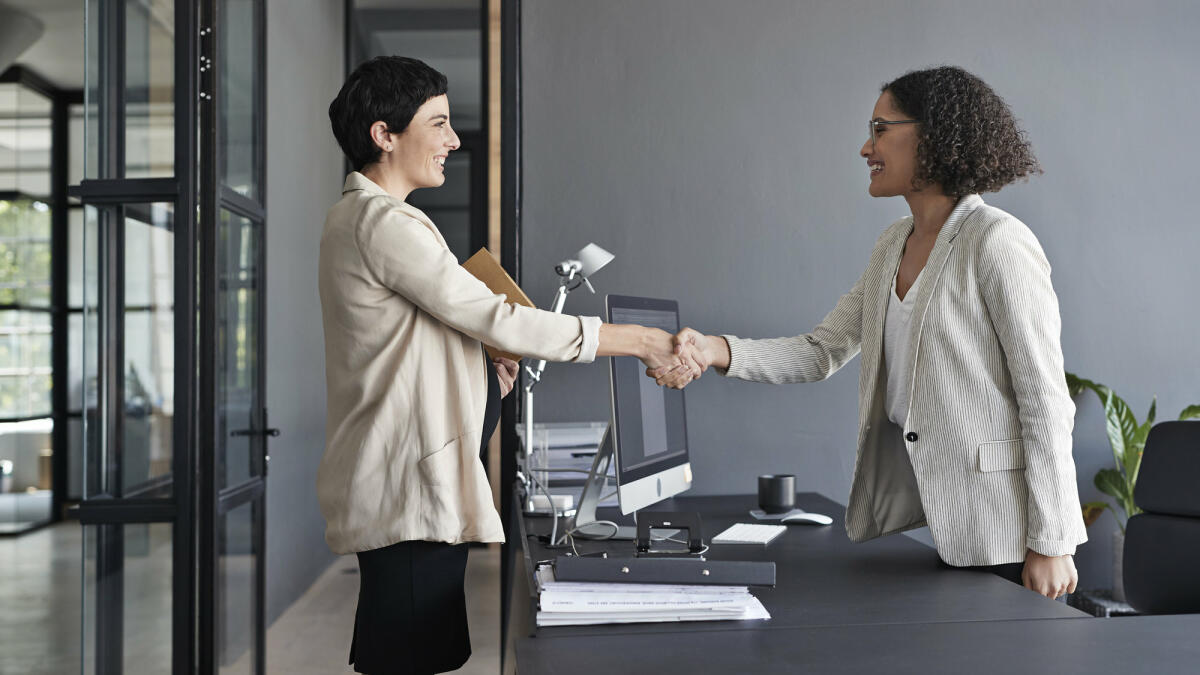Zwei Frauen geben sich im Büro über den Schreibtisch hinweg die Hand. © Klaus Vedfelt / Getty Images
