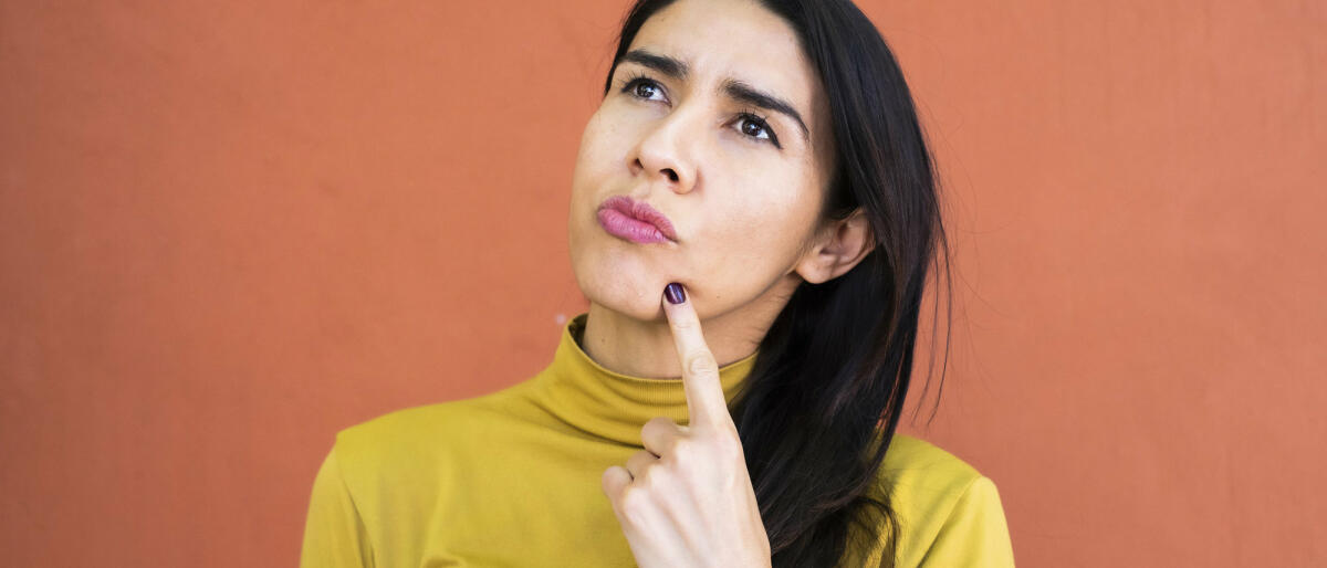 Latino-Frau in Gedanken versunken, nachdenklich und aufblickend, Finger am Kinn © Sergio Mendoza Hochmann / Getty Images
