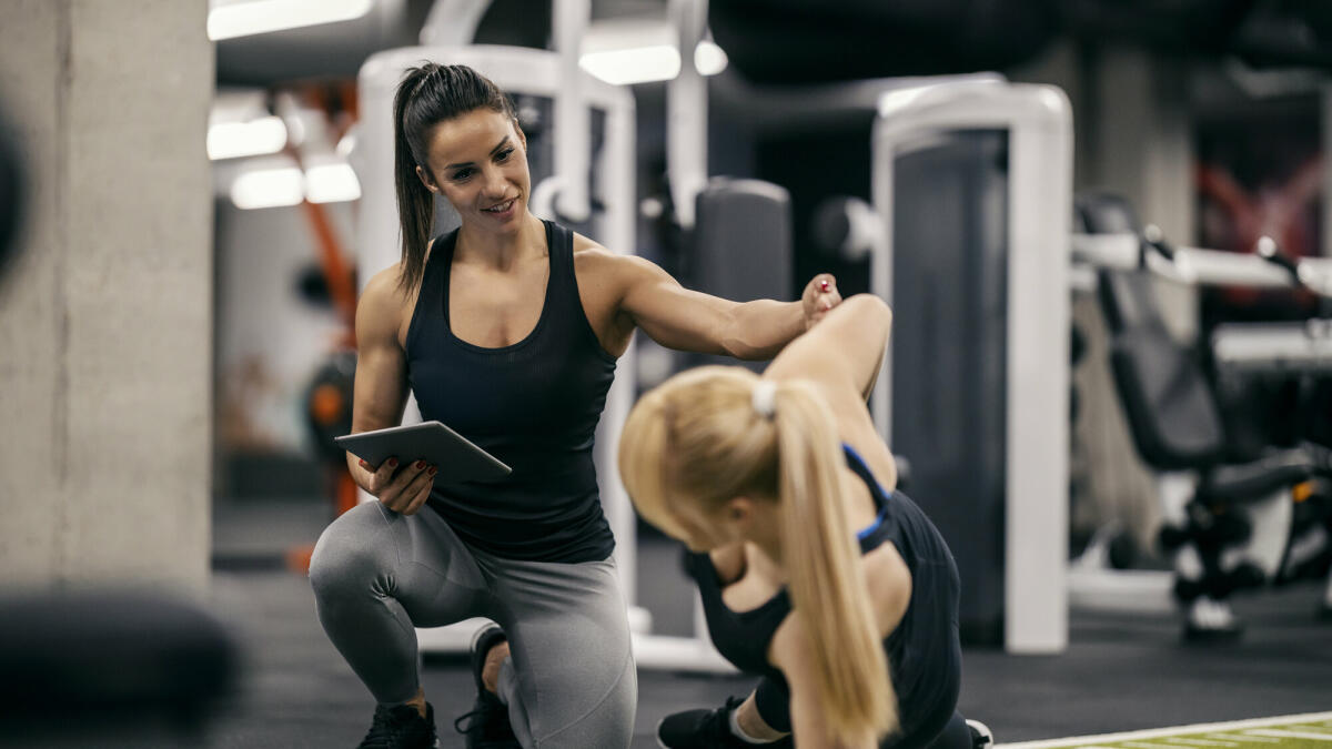 Eine Personal Trainerin trainiert eine Sportlerin in einem Fitnessstudio und verfolgt ihre Fortschritte auf einem Tablet. © dusanpetkovic/ Getty Images